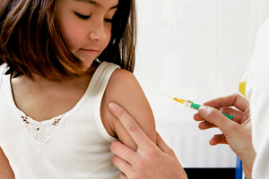 A preschool-aged girl in a tank top looks on as a health care provider gives her a shot in the upper arm