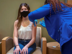 A teenage girl sits in a chair while a healthcare worker administers the coronavirus vaccine to her upper arm