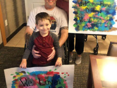 A white man in his 40s in a wheelchair. His wife is crouching next to him and holding a sign that says "Welcome Home Daddy! We missed you!" A young boy is standing in front of the man, holding a sign that says "We Love You!"