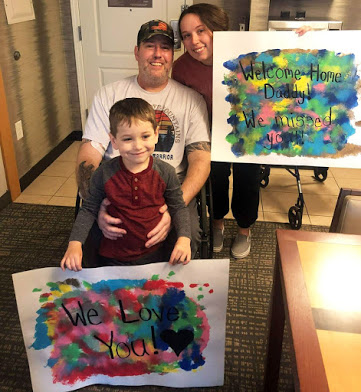 A white man in his 40s in a wheelchair. His wife is crouching next to him and holding a sign that says "Welcome Home Daddy! We missed you!" A young boy is standing in front of the man, holding a sign that says "We Love You!"