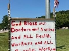 A large white sign on poles on grass in Jackson, Ky. with small American flags taped to both poles. The sign is written in red spray paint and says "God Bless our Doctors and Nurses and ALL Health Workers and ALL People Working