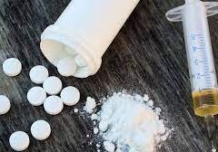 An open pill bottle with round white pills spilling out, next to a small pile of white powder and a syringe with a small amount of brown liquid in it.