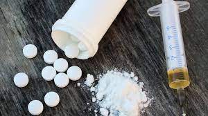 An open pill bottle with round white pills spilling out, next to a small pile of white powder and a syringe with a small amount of brown liquid in it.